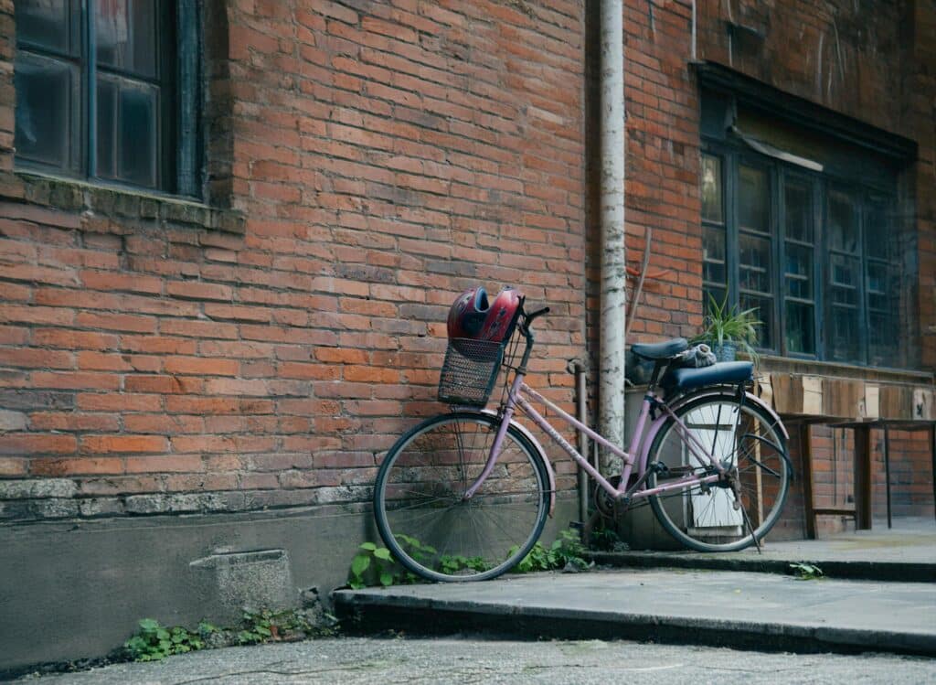 A pink bicycle with a basket and helmet leans against an urban brick wall.