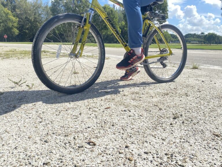 Bright yellow mountain bike on a gravel path with a rider wearing black and red athletic shoes and blue jeans.